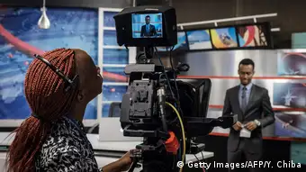 in a tv studio, a woman looks at the picture of a news anchor on a camara display 