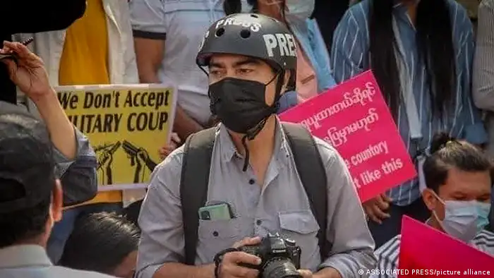 A reporter holding a photo camera wearing a helmet with the word press on it and a face mask is surrounded by protesters holding signs with written anti-coup slogans