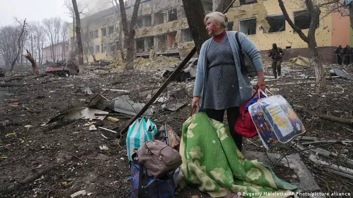 A woman stands with bags in front of a destroyed building