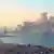 Внизу побережье, застроенное частными домами, далее синее море, небо без облаков. На воде слева поднимается дым от пожара на тонущем российском военном корабле "Орск". По центру два корабля, справа у берега небольшое судно.