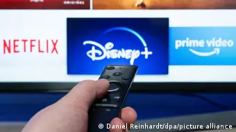 Netflix, Disney+ and Prime Video logos on a television, with a hand holding a remote control in the foreground