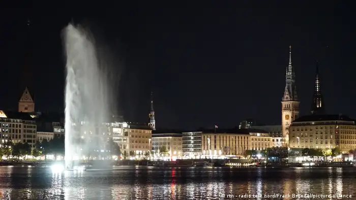 The large fountain spurts water with the city of hamburg illuminated in the background at night.