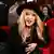 Alla Pugacheva wears a hat and looks at the camera while sitting on a plush red chair