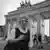 Schwarzweiß Foto: Eine Frau im Kleid mit grauen Haaren und Brille lacht freundlich in die Kamera, im Hintergrund ist das Brandenburger Tor zu sehen
