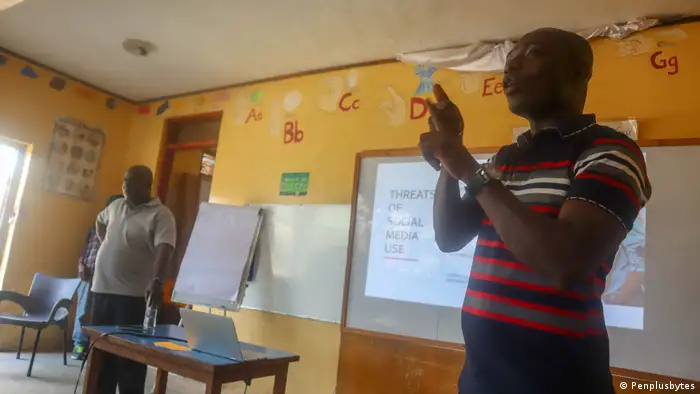 Dr. Kodwo Boateng (links, im Hintergrund) und Frederick Anderson stehen in einem Klassenraum. Anderson erklärt etwas in Gebärdensprache. Im Hintergrund wird eine Präsentation mit einem Beamer auf ein Whiteboard geworfen.