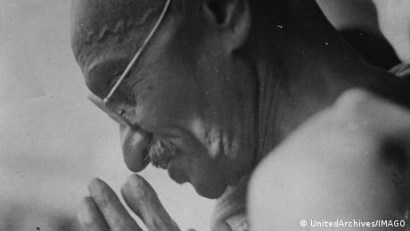A black and white close-up photograph of Mahatma Gandhi, his hands clasped in front of his nose