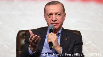 The picture shows Turkish President Tayyip Erdogan at an event in Bilecik. He is sitting and holding a micorphone, his right hand turned towards the audience. 