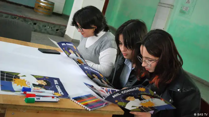 Three women sit at a desk reading a MIL comic book.