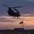 Helicóptero militar voa ao anoitecer, com bandeira dos EUA ao fundo 