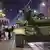 A tank draped in a little camouflage, with a soldier on top, at night in a city street. Other tanks in the background, and a small group of young civilians behind and to the left, one fliming on a phone.