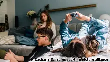 25/05/2023**ILLUSTRATION - Eine junge Frau liegt mit dem Ruecken auf der Couch am 25.05.2023 in Berlin, sie hat ihr Handy in beiden Haenden, ihre Geschwister sind auch im Wohnzimmer auf dem Sofa und mit ihren Social Media Kontakten beschaeftigt (gestellte Szene). Foto: Zacharie Scheurer || Modellfreigabe vorhanden || Modellfreigabe vorhanden