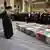 Aiatolá Ali Khamenei de pé, olhando para caixões cobertos com bandeiras do Irã e fotos das pessoas mortas