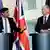 El primer ministro británico habla desde el atril dirigiéndose al canciller alemán, que le escucha desde el atril contiguo, con las banderas de la Unión Europea, el Reino Unido y Alemania detrás.